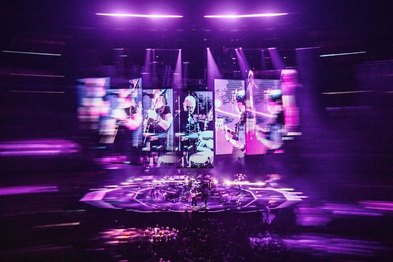 Wide-angle live concert shot of Radiohead performing on stage beneath purple lighting and large visual screens.